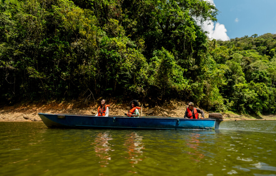 A group of people wearing life jackets enjoy a scenic boat ride along a calm river, surrounded by dense green forest and blue skies.