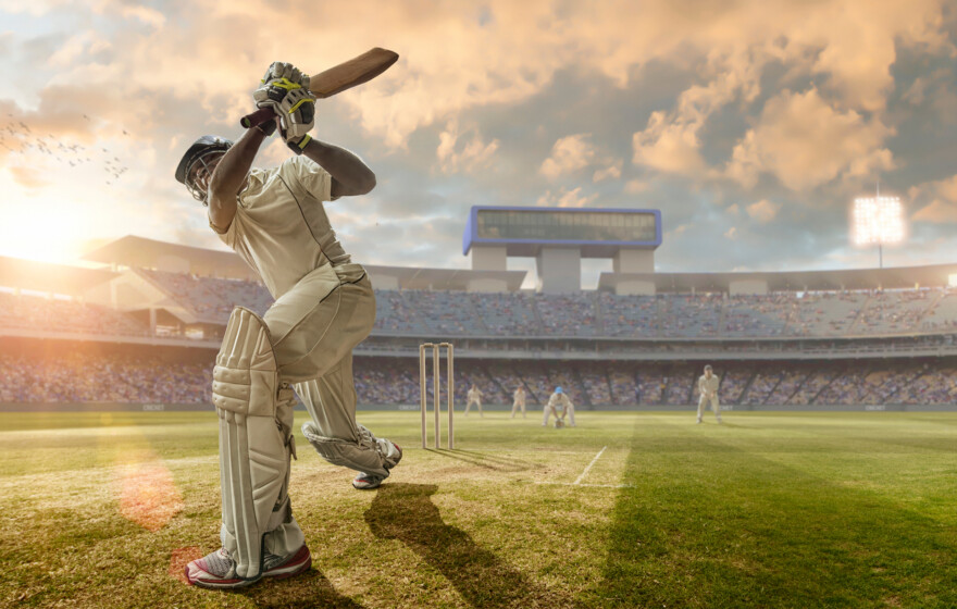 A close up image of a professional cricketer playing in batsman position wearing cricket whites and safety helmet, having just hit a ball during a cricket match in an outdoor stadium full of spectators. The action occurs under an evening sky at sunset. Stadium is fake, created from photographic and CG elements.